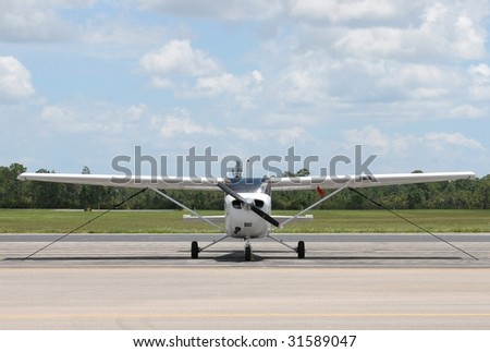 A small, overhead wing private aircraft tied down on a concrete ramp at an airport in a remote location. The view is from straight ahead. It is a sunny day. The blue sky has small, puffy white clouds