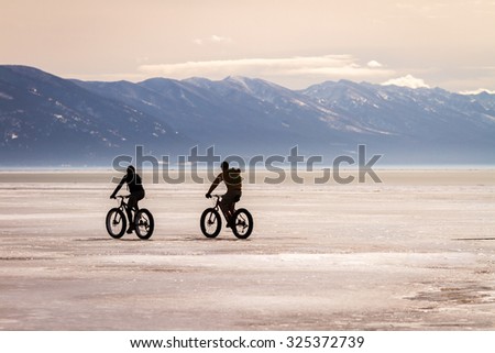 Two bicyclists ride fat tire bikes across a frozen Flathead lake with the Mission Mountain range in the background near Kalispell MT.