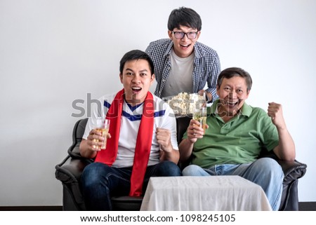 Young Happy Asian Men family or football fans watching soccer match on tv and cheering football team, celebrating with drink beer and eat popcorn at home, sports and entertainment concept.