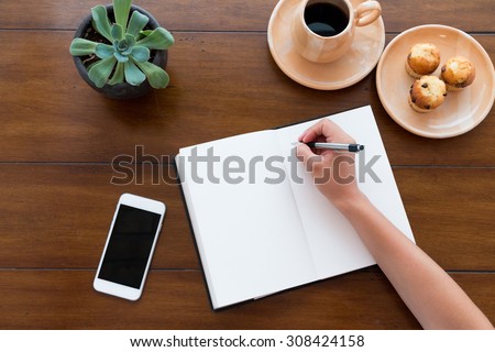 A person writing on her white notebook next to her smarth phone on a beautiful new wooden table with coffee and muffins