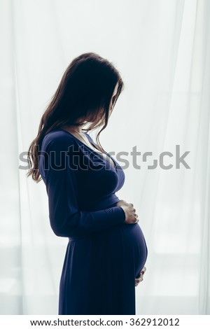 Silhouette of pregnant woman in a pink negligee standing against the window. Side view of happy pregnant woman looking at belly