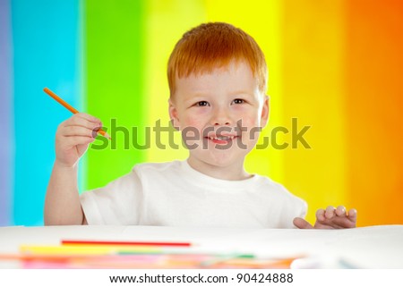 Red-haired adorable boy drawing with orange pencil on rainbow background