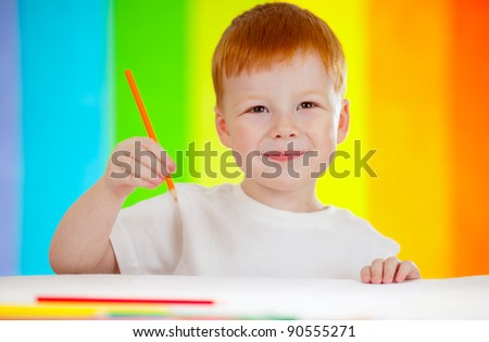 Red-haired adorable boy drawing with orange pencil on rainbow background