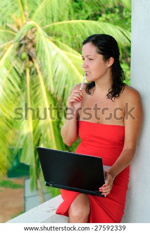 Pondering beautiful young woman in red dress with laptop on knee typing