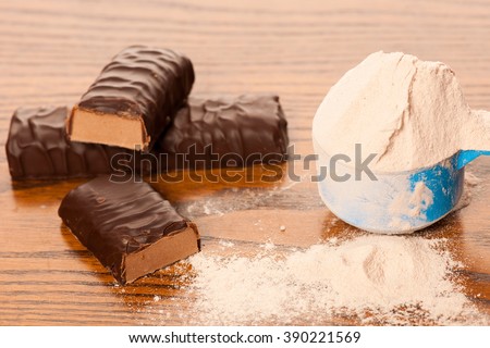 Whey protein powder in measuring scoop and chocolate protein bar on wooden background.