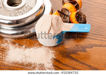 Whey protein powder in measuring scoop, meter tape and dumbbell on wooden background.