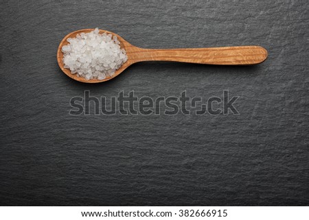 white crystals sea salt on wooden spoon on black stone background, top view