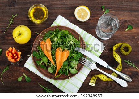 The concept of diet food. Salad with arugula and carrots in a clay plate. Beside the yellow sewing tape and the ingredients for salads, like as carrot, arugula, leaf mash, olive oil