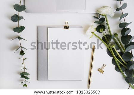 Eucalyptus and ranunculus flower, laptop keyboard, pen, grey copybook with paper mock-up on white table top view. Plants in home office. Feminine floral desktop.