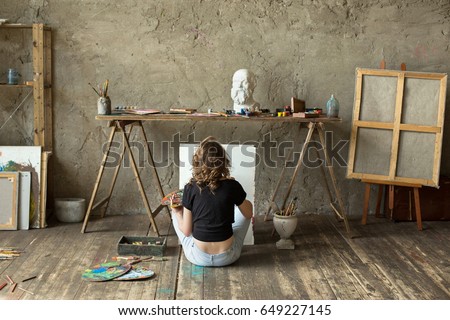 Woman painter sitting on the floor in front of an empty canvas and drawing. Art studio interior. Horizontal background. Drawing supplies, oil paints, artist brushes, canvas, frame. Creative concept