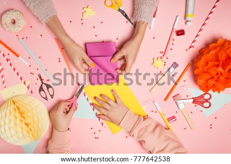 Colorful pink background with various party confetti, paper decoration, flags, stationary, DIY accessories with woman\'s and kid\'s hands making greeting card. Fat lay top view. Party arrangement