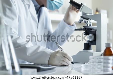 Scientist conducting research taking notes whilst looking through microscope selective focus