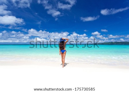 Beautiful woman on white sandy beach,  holding her hat and looking to sea.  Back view of slim girl in blue bikini.