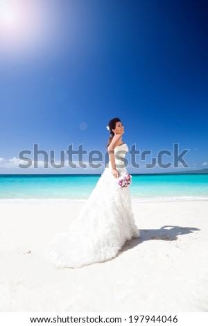 Beautiful bride in wedding gown with train, on white sandy beach, smiling and feeling happiness. Travel wedding concept.