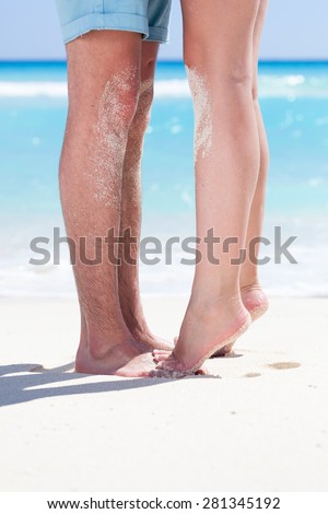 Woman\'s barefoot legs standing up tiptoe closeup to male foot and kissing on sandy beach with turquoise sea background, no face. Happy romantic holidays concept.