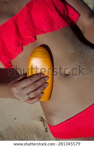 Tan female belly on sand with sunscreen cream bottle in hand on it, close up, no face. Sunblock protection for healthy skin on tropical vacation.