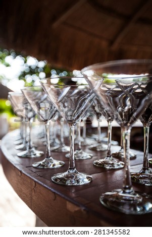 Empty Martini glasses setting in row, outside on wooden bar counter at tropical grass palapa,  ready for celebration party, selective focus