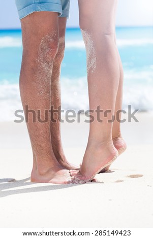 Woman\'s barefoot legs standing up tiptoe closeup to male foot and kissing on sandy beach with turquoise sea background, no face. Happy romantic holidays concept.