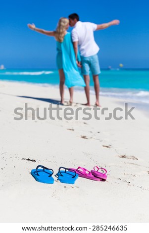 Beach color flip-flops on sandy seashore closeup on background with happy man and woman, enjoying summer vacation, kissing with outstretched arms, back view. Travel concept