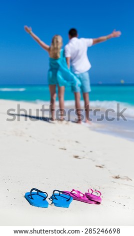 Beach color flip-flops on sandy seashore closeup on background with happy man and woman, enjoying summer vacation, looking to the sea with outstretched arms, back view. Travel concept