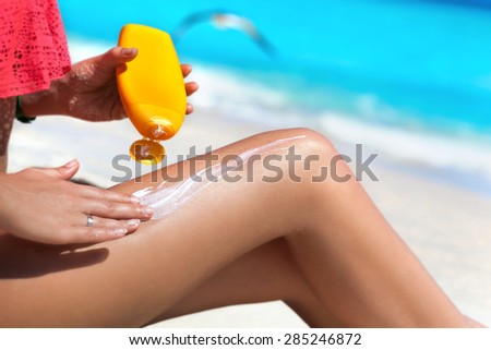 Tan slim woman applying sunscreen on her legs, sitting on sandy beach with sea background. SPF sunblock protection concept. Travel vacation
