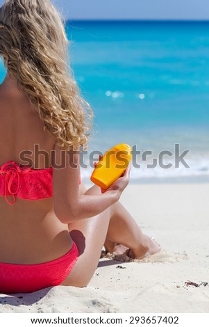 Blond tan woman on summer vacation is sitting on beach near turquoise sea and holding sunscreen lotion in hand, back view, no face, focus on cream tube. Healthy tanning concept.