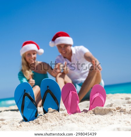 Caribbean Christmas vacation. Two pair of flip flops standing in a sand on background with  happy couple in Santa  hats, sitting on beach, smiling and showing thumbs up. Focus on sandals