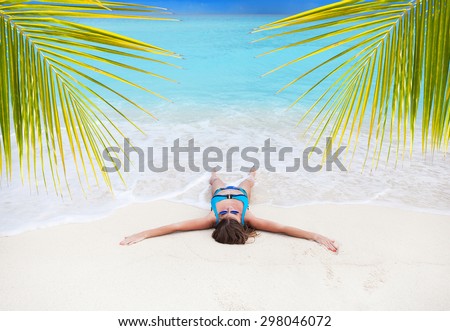 Beautiful tan woman in bikini lying like a sea star on white sandy beach near turquoise sea through palm tree leafs