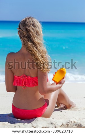 Blond tan woman on summer vacation is sitting on beach near turquoise sea and holding sunscreen lotion in hand, back view, no face, focus on cream tube. Healthy tanning concept.