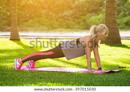 Young beautiful fitness woman with ponytail doing plank position outside on the green grass at the park