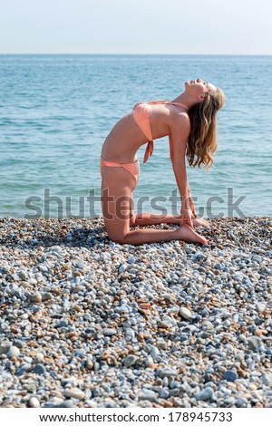 Supple young woman exercising on the beach kneeling down on the pebbles arching back gracefully to touch her toes in a health and fitness concept