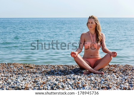Beautiful woman practicing yoga meditating at the beach sitting cross-legged in the lotus position on the sand with the ocean behind her and copyspace