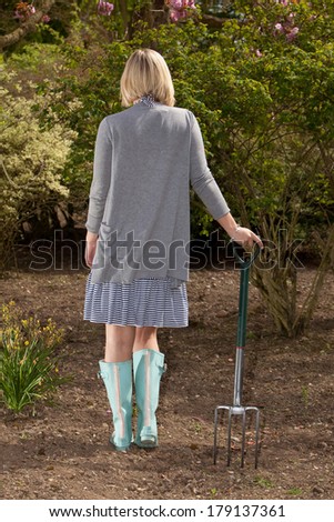 Woman gardener holding a metal fork standing in a dress, jersey and gumboots with her back to the camera in a shaft of sunlight