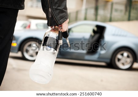 Drunk man driver entering a car with a bottle of alcohol in his hand, his car in the background