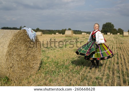 A pretty young smiling girl dressed in Polish national folk dress dancing and spinning around in the field of harvest straw bales