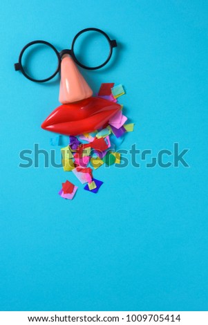 high angle view of a pair of fake black glasses, a nose and a mouth forming the face of a person, and a pile of confetti on a blue background, with a blank space on the bottom