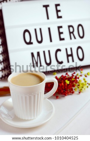 closeup of a white ceramic cup with coffee on a table, a bunch of flowers and a lightbox in the background with the text te quiero mucho, I love you so much written in spanish