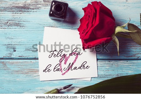 a red rose, an ink bottle, a nib pen and a piece of paper with the text feliz dia de la madre, happy mothers day written in spanish, on a blue rustic wooden table