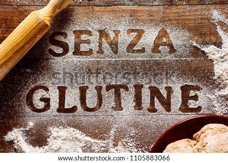 high angle view of a wooden table sprinkled with a gluten free flour where you can read the text senza glutine, gluten free in italian, next to a rolling pin and a piece of dough