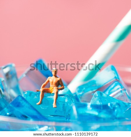 a miniature man wearing swimsuit and swim cap relaxing on the ice cubes of a blue cocktail served in a cocktail glass, against a pink background with some blank space on top