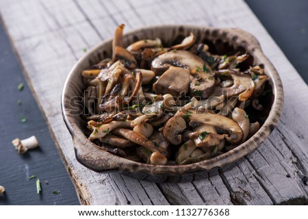 closeup of a brown earthenware bowl with some cooked mixed mushrooms, such as common mushrooms, oyster mushrooms or shiitake, on a white rustic wooden board