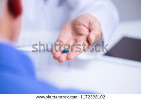 closeup of a caucasian doctor man, in a white coat, giving a blue pill to a senior caucasian patient man, sitting both at a doctors desk