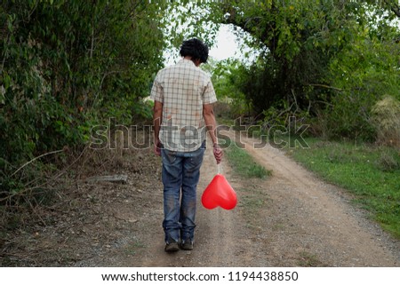 a scary man wearing dirty and ragged clothes, seen from behind, with a red heart-shaped balloon walking by a dirt road in a disturbing landscape