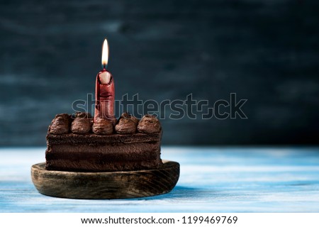 closeup of a multilayered chocolate cake topped with a bloody middle finger, with a lit wick on top, on a blue rustic wooden table, with some blank space on the right