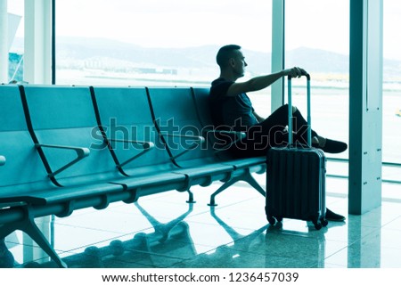 a young caucasian man, with his trolley case, waiting for his flight sitting at the waiting room of an airport