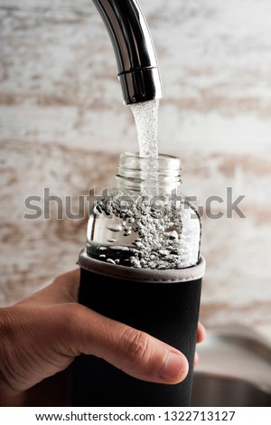 closeup of a young cacausian man filling a glass reusable water bottle with water from a tap of a filtered water station or a domestic kitchen