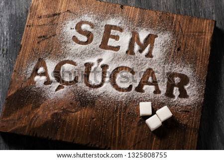 high angle view of a wooden chopping board sprinkled with sugar where you can read the text sem acucar, sugar free written in Portuguese, and some sugar cubes