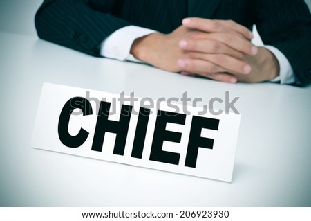 a man wearing a suit sitting in a desk with a signboard in front of him with the word chief written in it
