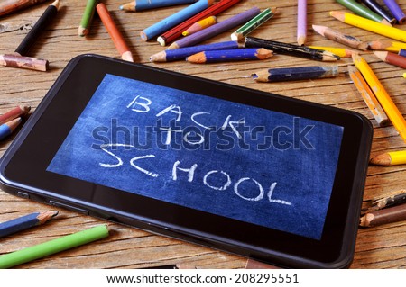 a tablet with a picture of a chalkboard with the sentence back to school written in it, on a rustic wooden desk with worn pencil crayons of different colors