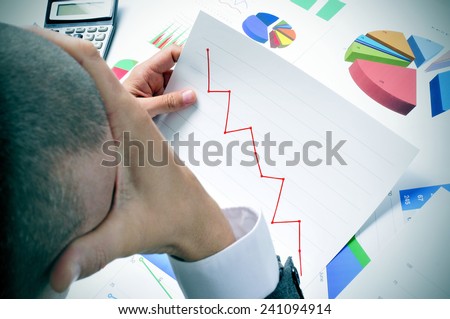 businessman in his office desk full of graphs and charts observing worried a chart with a downward trend